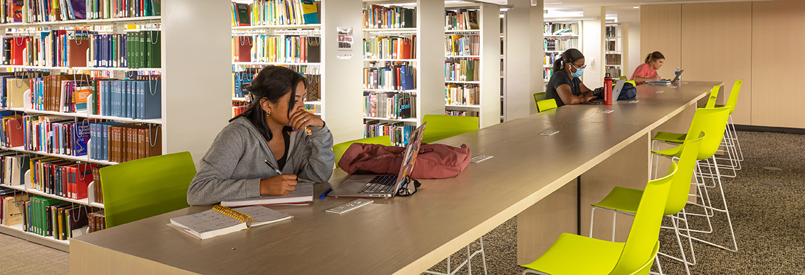 student study at table sitting at brightly colored chairs in front of shelves of books