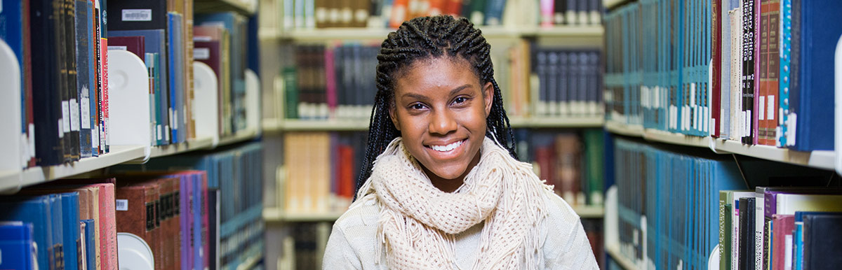young student standing among library stacks smiling at camera